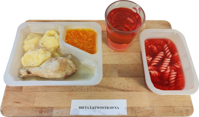 Na drewnianej tacy widoczny jest zestaw obiadowy z kartką "Dieta łatwostrawna". W podzielonym pojemniku znajdują się: puree ziemniaczane z sosem, pieczone udko z kurczaka oraz surówka z marchwi. Obok stoi szklanka z czerwonym napojem oraz osobny pojemnik z makaronem w czerwonej zupie