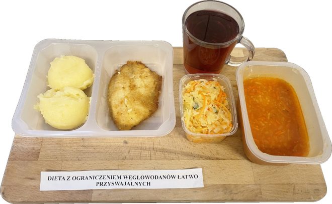 Na drewnianej tacy widoczny jest zestaw obiadowy z kartką "Dieta z ograniczeniem węglowodanów łatwo przyswajalnych". W pojemniku znajduje się puree ziemniaczane, filet z ryby w panierce oraz surówka z kapusty i marchewki. Obok w przezroczystym pojemniku podano zupę pomidorową. Na tacy stoi również szklanka z herbatą