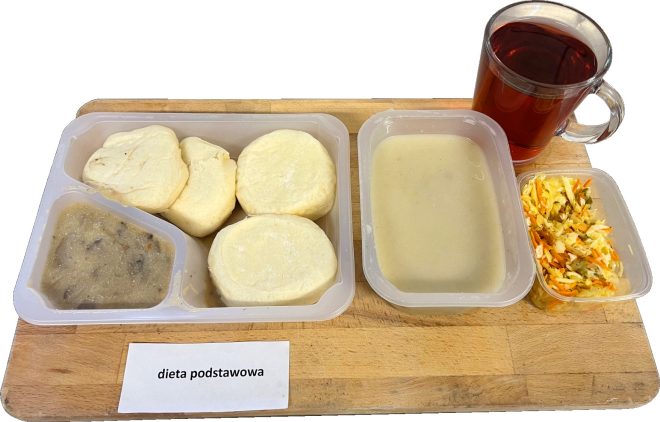 Na drewnianej tacy ustawiono zestaw obiadowy oznaczony jako "Dieta podstawowa". W plastikowym talerzu znajdują się cztery knedle, sos pieczarkowy w oddzielnym przegródce oraz jasny sos. Obok podano porcję surówki z kapusty, marchwi i ogórka kiszonego w plastikowym pojemniku oraz kubek herbaty w przezroczystym szkle