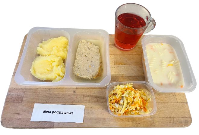 Na drewnianej desce ustawiono zestaw posiłku oznaczonego jako „Dieta podstawowa”. W plastikowych pojemnikach znajduje się porcja puree ziemniaczanego, kawałek mięsa w sosie oraz zupa z makaronem. Obok mały pojemnik z surówką z marchewki i kapusty oraz szklanka czerwonego kompotu