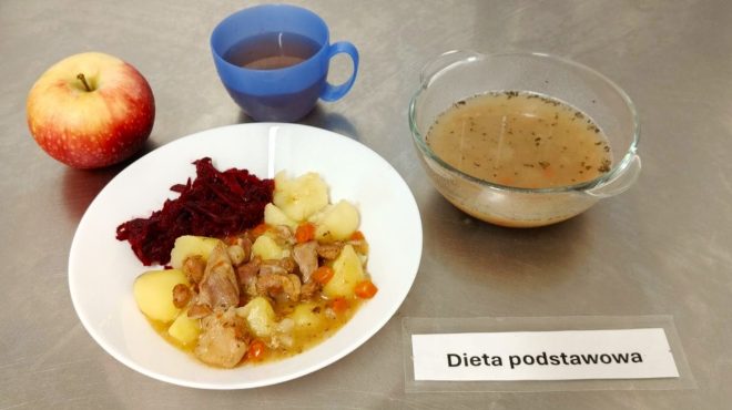 Zestaw obiadowy „Dieta podstawowa”. Na talerzu znajduje się gulasz z ziemniakami i marchewką, porcja buraczków na ciepło oraz jabłko. Obok szklanka herbaty i miseczka z zupą jarzynową, posypaną ziołami