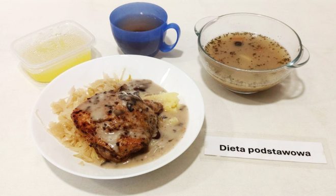 Na talerzu widoczna jest porcja kotleta wieprzowego z sosem pieczarkowym, podana z kapustą zasmażaną i ziemniakami puree. Obok ustawiono filiżankę z herbatą, przezroczystą miseczkę z rosołem oraz plastikowy pojemnik z kompotem. Na stole umieszczono kartkę z napisem "Dieta podstawowa", określającą rodzaj posiłku