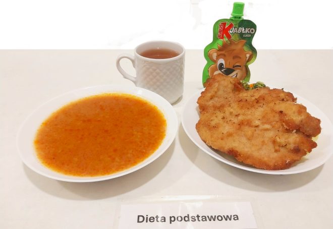 Na białym tle przedstawiono zestaw obiadowy opisany kartką „Dieta podstawowa”. Na talerzu znajduje się panierowany kotlet schabowy oraz porcja surówki warzywnej. Obok na drugim talerzu znajduje się talerz zupy pomidorowej, a całość uzupełnia kubek herbaty i owocowy mus w opakowaniu typu „Kubuś”