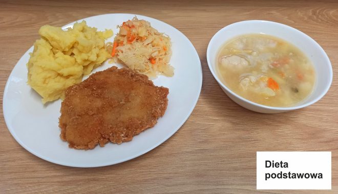 Na talerzu zestaw posiłku oznaczonego jako „Dieta podstawowa”. Danie główne to panierowany kotlet schabowy, porcja puree ziemniaczanego oraz surówka z kapusty kiszonej i marchewki. Obok talerza ustawiona jest miska z rosołem zawierającym kawałki warzyw i mięsa