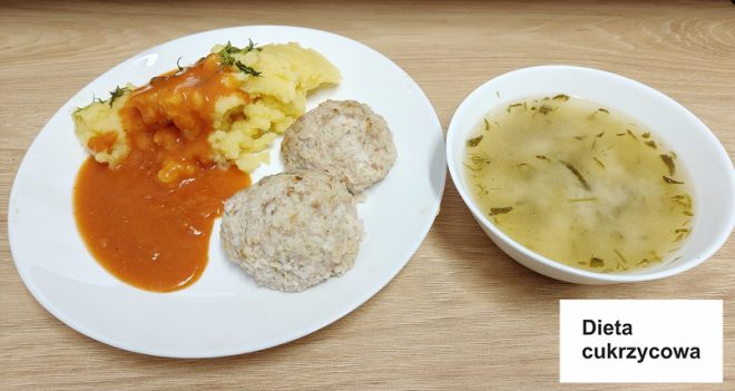 Na drewnianym blacie widoczny jest zestaw obiadowy. Na białym talerzu podane są dwie pulchne, gotowane pulpety z mięsa mielonego, porcja puree ziemniaczanego polana sosem pomidorowym i posypana świeżym koperkiem. Obok, w białej miseczce, znajduje się klarowna zupa z kawałkami ziół i warzyw. Obok umieszczona jest karteczka z napisem „Dieta cukrzycowa”