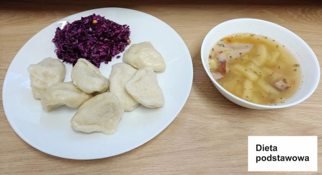 Zdjęcie przedstawia obiad w ramach diety podstawowej. Na białym talerzu ułożone są pierogi z nadzieniem, a obok nich znajduje się porcja surówki z czerwonej kapusty. Obok talerza ustawiona jest miseczka z zupą jarzynową zawierającą kawałki ziemniaków, marchewki i mięsa, przyprawioną ziołami. Posiłek podany jest na drewnianym blacie, a w prawym dolnym rogu zdjęcia widnieje napis „Dieta podstawowa”