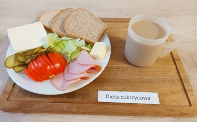 Na drewnianej desce umieszczono biały talerz z elementami śniadania. Na talerzu znajdują się trzy kromki chleba pełnoziarnistego, plasterki pomidora, ogórka kiszonego, kawałki sałaty, plasterki wędliny, kostka białego sera oraz mały kawałek masła. Obok postawiono plastikowy kubek z jasnym napojem (prawdopodobnie kawa z mlekiem). Na desce widnieje kartka z napisem „Dieta cukrzycowa”