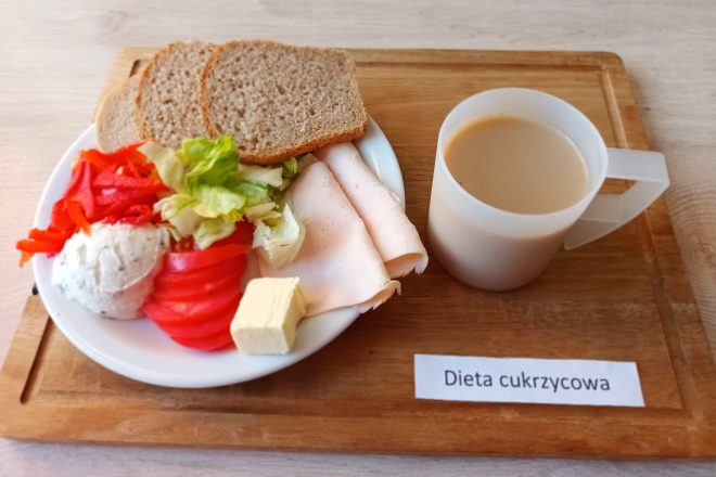 Na drewnianej tacce zaprezentowano zestaw śniadaniowy przeznaczony dla diety cukrzycowej, co zostało oznaczone kartką z napisem „Dieta cukrzycowa”. Na talerzu znajdują się plasterki wędliny drobiowej, sałatka z liści sałaty, kawałków papryki i plasterków pomidora, a także serek twarogowy z ziołami i kostka masła. Towarzyszą im dwie kromki pełnoziarnistego chleba. Obok talerza stoi kubek z kawą z mlekiem