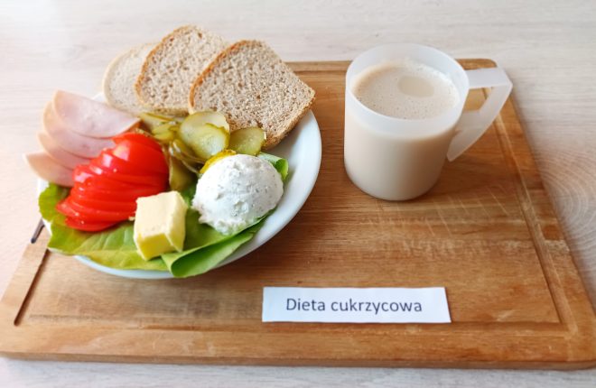 Na drewnianej tacy znajduje się zestaw śniadaniowy oznaczony jako "Dieta cukrzycowa". Na białym talerzu podano dwie kromki pieczywa pełnoziarnistego, kilka plasterków szynki, kawałki pomidora i ogórka konserwowego, kostkę masła oraz porcję twarożku z ziołami na liściu sałaty. Obok stoi kubek z napojem mlecznym, prawdopodobnie kawą z mlekiem lub mlekiem bez dodatków