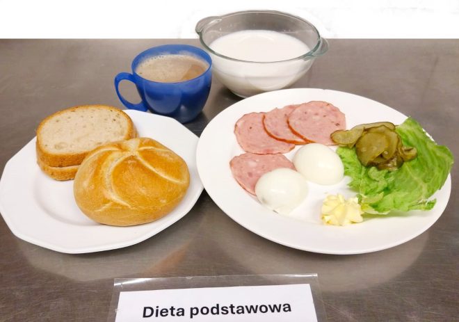 Zestaw śniadaniowy oznaczony kartką „Dieta podstawowa”. Na białych talerzach podano dwa rodzaje pieczywa: kromkę chleba jasnego, kromkę chleba ciemnego oraz bułkę. Towarzyszy im talerz z plasterkami szynki drobiowej, dwoma jajkami na twardo, plasterkami ogórka kiszonego, liściem sałaty i kostką masła. Obok znajduje się miseczka z mlekiem oraz kubek z kawą