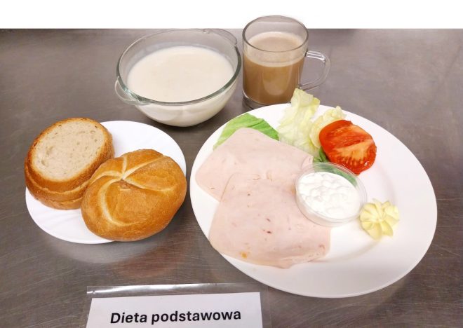 Zestaw śniadaniowy przedstawiony na srebrnym blacie i opisany kartką „Dieta podstawowa”. Na jednym talerzu znajdują się kromki chleba jasnego i ciemnego oraz bułka, a na drugim plasterki wędliny drobiowej, plasterki pomidora, liść sałaty, mała porcja twarogu oraz kostka masła. Całość dopełniają miska z mlekiem i kubek z kawą