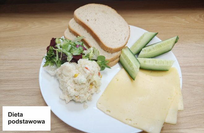 Kolacja dla diety podstawowej na jasnym blacie. Na białym talerzu umieszczono kilka kromek pieczywa, plastry żółtego sera, kawałki świeżego ogórka, porcję sałatki jarzynowej oraz mieszankę liści sałat z dodatkiem rukoli. Całość wygląda świeżo i estetycznie. Na białym tle widoczny jest napis "Dieta podstawowa"