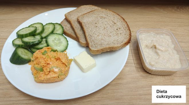 Na białym talerzu podano trzy kromki pełnoziarnistego chleba, porcję pasty warzywnej o pomarańczowym odcieniu, dekorowanej posiekanym szczypiorkiem, oraz kilka plasterków świeżego zielonego ogórka. Obok na talerzu znajduje się mały kawałek masła. Na blacie, obok talerza, stoi przezroczysty pojemnik z jasnobeżową pastą, prawdopodobnie twarożkiem lub pasztetem. Całość ułożona jest na jasnym drewnianym blacie. W prawym dolnym rogu zdjęcia widnieje etykieta z napisem „Dieta cukrzycowa” na białym tle