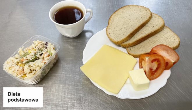 Na metalowym blacie znajduje się śniadanie oznaczone jako „Dieta podstawowa”. Na białym talerzu ułożono trzy kromki jasnego chleba, dwa plastry żółtego sera, plasterki pomidora oraz małą kostkę masła. Obok talerza stoi przezroczysty pojemnik z sałatką ryżową zawierającą kawałki warzyw i mięsa. W tle biała filiżanka z ciemnym napojem, prawdopodobnie herbatą