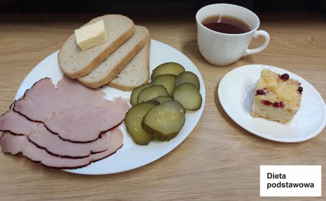 Na zdjęciu znajduje się kolacja składająca się z białego talerza z różnorodnymi produktami spożywczymi. Na talerzu leżą dwie kromki jasnego chleba pszennego, jedna z kawałkiem masła, kilka plastrów wędzonej szynki oraz plasterki ogórków kiszonych. Obok, na małym białym talerzyku, znajduje się kawałek ciasta drożdżowego z żurawiną. Do posiłku podano również filiżankę herbaty w białej porcelanowej filiżance. Posiłek jest ułożony na drewnianym blacie, a w prawym dolnym rogu zdjęcia umieszczona jest etykieta z napisem "Dieta podstawowa"