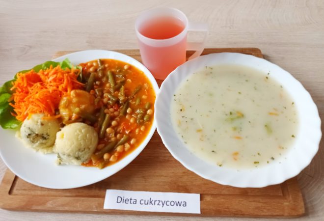 Zdjęcie przedstawia posiłek oznaczony jako „Dieta cukrzycowa”. Na drewnianej tacy znajduje się biały talerz z dwoma kulkami ziemniaków puree, ciepłymi warzywami w sosie pomidorowym (zielona fasolka, biała fasola, marchewka) oraz startą marchewką na liściu sałaty. Obok stoi miska z zupą jarzynową w jasnym bulionie i przezroczysty kubek z herbatą owocową