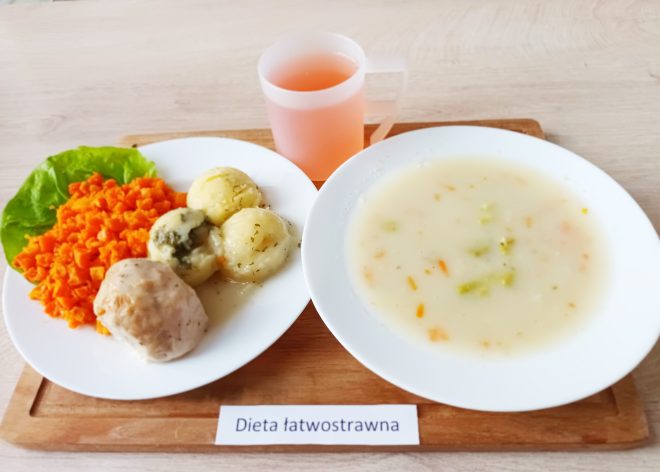 Na drewnianej tacy podano zestaw obiadowy w ramach diety łatwostrawnej. Na białym talerzu znajduje się porcja drobno posiekanej marchewki, puree ziemniaczane polane lekkim sosem oraz kawałek gotowanego mięsa w jasnym sosie. Obok znajduje się miseczka z jasną, kremową zupą warzywną, w której widać kawałki marchewki i zielonego warzywa. Do posiłku podano także kubek z różowym napojem, najprawdopodobniej kompotem. Na drewnianej tacy umieszczono karteczkę z napisem "Dieta łatwostrawna"