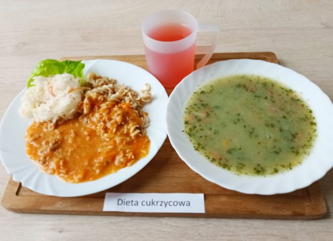 Na drewnianej tacy znajduje się zestaw obiadowy oznaczony jako „Dieta cukrzycowa”. Na białym talerzu podano porcję gęstej zupy jarzynowej o zielonkawym odcieniu, z widocznymi kawałkami marchwi, ziemniaków i posiekaną natką pietruszki. Obok, na drugim talerzu, znajduje się porcja pełnoziarnistego makaronu z gulaszem mięsnym w pomarańczowym sosie, obok surówka z kiszonej kapusty udekorowana liściem sałaty. Do posiłku podano różowy napój w plastikowym kubku. Całość ustawiona jest na jasnym blacie stołu