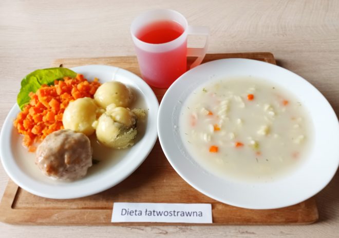 Na drewnianej tacy znajduje się zestaw posiłków przeznaczonych dla osoby na diecie łatwostrawnej. Na białym talerzu umieszczono trzy ziemniaki polane lekkim sosem, mielony kotlet w sosie oraz gotowaną marchewkę pokrojoną w drobną kostkę. Obok stoi kubek z przezroczystą czerwoną herbatą. Na drugim talerzu znajduje się jasna zupa z makaronem i warzywami – kawałkami marchewki i koperkiem. Całość wygląda na delikatny i lekkostrawny posiłek. Na desce znajduje się karteczka z napisem „Dieta łatwostrawna”