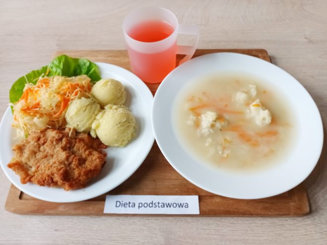 Na zdjęciu widoczny jest obiad podany na białym talerzu z dodatkami. W zestawie znajduje się panierowany kotlet schabowy, trzy porcje puree ziemniaczanego udekorowane koperkiem, surówka z kapusty kiszonej z marchewką, jasna zupa kalafiorowa z kawałkami marchewki podana na osobnym talerzu oraz kubek różowego kompotu. Etykieta informuje, że to „Dieta podstawowa”