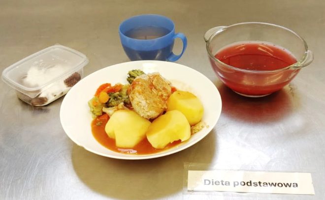 Na zdjęciu widzimy talerz obiadowy oznaczony jako "Dieta podstawowa". Na talerzu znajduje się porcja gotowanych ziemniaków, pieczeń wieprzowa w sosie pomidorowym, a także warzywa na parze (brokuły, marchewka, papryka). Obok talerza umieszczono plastikowy pojemnik z deserem, kubek niebieskiego koloru z herbatą oraz szklane naczynie z kompotem owocowym