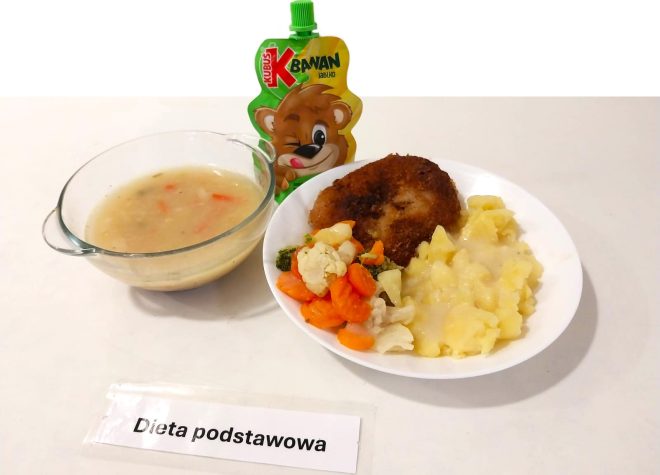 Na zdjęciu widoczny jest zestaw obiadowy oznaczony jako „Dieta podstawowa”. Na białym talerzu znajduje się kotlet mielony, porcja ziemniaków polanych jasnym sosem oraz warzywa gotowane, takie jak marchewka, brokuły i kalafior. Obok ustawiono miseczkę z zupą warzywną oraz opakowanie musu owocowego „Kubuś” o smaku jabłkowym