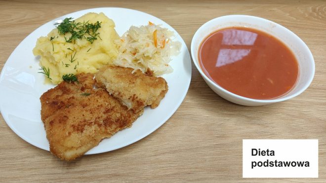 Na białym talerzu znajduje się panierowany filet rybny o złocistej, chrupiącej skórce, obok niego porcja gładkiego puree ziemniaczanego, posypanego świeżym koperkiem. Dopełnieniem dania jest surówka z kiszonej kapusty z dodatkiem marchewki. Obok talerza, w białej miseczce, znajduje się talerz zupy pomidorowej o głębokim czerwonym kolorze. Całość ustawiona jest na jasnym drewnianym blacie. W prawym dolnym rogu zdjęcia znajduje się etykieta z napisem „Dieta podstawowa” na białym tle