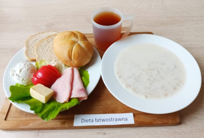 Zdjęcie prezentuje posiłek oznaczony jako „Dieta łatwostrawna”. Na drewnianej tacy znajduje się talerz z bułką pszenną, dwoma kromkami jasnego chleba, plastrami wędliny, kawałkiem masła, połową pomidora, gałką twarożku i liściem sałaty. Obok znajduje się miska z zupą mleczną i przezroczysty kubek z herbatą