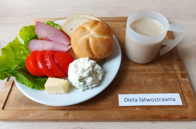 Dieta łatwostrawna. Na drewnianej desce umieszczono biały talerz z kajzerką, kromką chleba, plastrem wędliny, plasterkami pomidora, kawałkiem masła, porcją twarogu i liściem sałaty. Obok stoi biały kubek z jasnym napojem, prawdopodobnie kawą zbożową lub mlekiem.