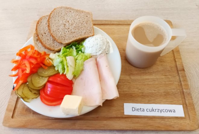 Na drewnianej tacy podano śniadanie w postaci białego talerza z różnorodnymi składnikami. Na talerzu znajdują się dwie kromki razowego chleba, dwa plastry chudej szynki drobiowej, liście sałaty, pokrojone w plastry pomidory, ogórki kiszone oraz czerwona papryka. Dodatkowo jest porcja serka twarogowego z ziołami i kawałek masła. Obok talerza stoi przezroczysty kubek z kawą z mlekiem. Na tacy umieszczono kartkę z napisem „Dieta cukrzycowa”