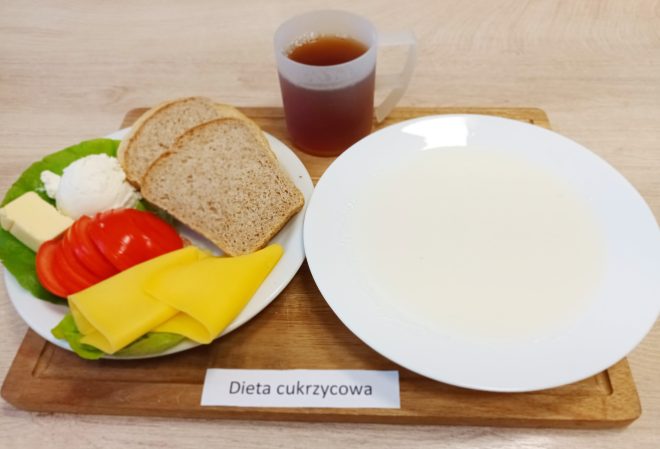 Na drewnianej tacy ustawionej na jasnym blacie znajduje się posiłek oznaczony jako „Dieta cukrzycowa”. Na białym talerzu ułożono dwa kromki razowego chleba, kilka plastrów żółtego sera, plasterki pomidora, porcję białego twarożku, kostkę masła oraz liść sałaty. Obok, na oddzielnym białym talerzu, znajduje się jasna zupa mleczna. W tle stoi przezroczysty plastikowy kubek z ciepłym napojem o bursztynowym odcieniu