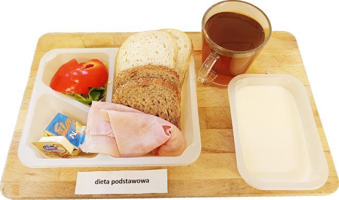 Na drewnianej tacy znajduje się śniadanie w plastikowych pojemnikach, oznaczone jako „DIETA PODSTAWOWA”. W zestawie są trzy kromki pieczywa (białe i pełnoziarniste), plasterki szynki, masło i serek topiony, a także porcje sałaty i czerwonej papryki. Obok znajduje się kubek ciemnego napoju oraz plastikowy pojemnik z białą, kremową zupą