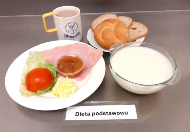 Na zdjęciu widoczny jest zestaw śniadaniowy opisany jako „Dieta podstawowa”. Na białym talerzu znajduje się plasterek pomidora, liść sałaty, szynka drobiowa, porcja dżemu oraz masło. Obok ustawiono miseczkę z mlekiem, pieczywo w postaci bułki pszennej i kromek chleba, a także kubek z napisem „Kryniczanka” zawierający gorący napój