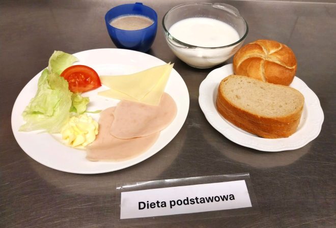 Na zdjęciu widoczny jest zestaw śniadaniowy oznaczony jako „Dieta podstawowa”. Na białym talerzu podano plasterki szynki drobiowej, liść sałaty, plasterek pomidora, plaster sera żółtego oraz niewielką porcję masła. Obok znajduje się miseczka z mlekiem, bułka pszenna i kromki chleba żytniego, a także kubek kawy z mlekiem w niebieskim naczyniu