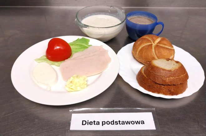 Na zdjęciu zaprezentowano śniadanie oznaczone jako „Dieta podstawowa”. Na białym talerzu znajduje się plasterek pomidora, sałata, kawałek mozzarelli, plasterki szynki drobiowej oraz masło. Obok ustawiono miseczkę z mlekiem, bułkę pszenną i kromki chleba razowego, a także kubek kawy z mlekiem w niebieskim naczyniu