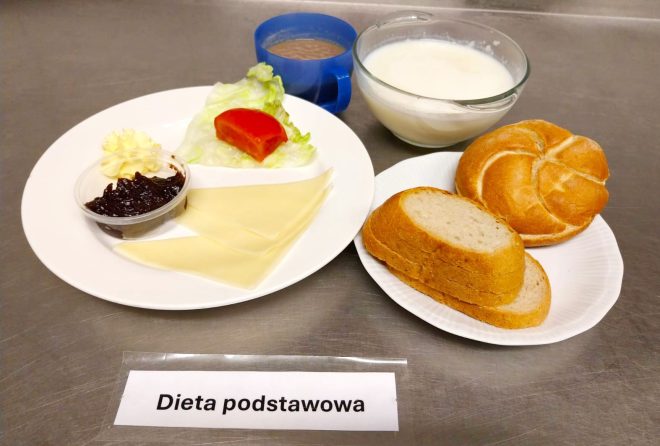 Zdjęcie przedstawia śniadanie oznaczone jako „Dieta podstawowa”. Na białym talerzu podano liść sałaty z plasterkiem pomidora, dwie plastry sera żółtego, niewielką porcję masła oraz dżem w małym pojemniku. Obok na talerzyku znajdują się bułka pszenna i kilka kromek chleba, a w naczyniach mleko oraz kawa z mlekiem w niebieskim kubku