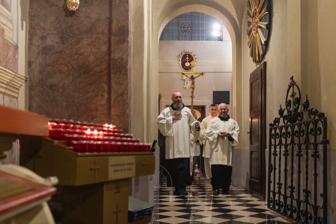 Procesja liturgiczna w bocznym korytarzu kościoła. Na czele duchowny z brodą trzymający duży złoty krzyż. W tle widoczne palące się świece oraz żelazna brama prowadząca do głównej nawy