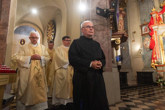 Duchowni w złotych ornatach kroczą w procesji przez świątynię. Przed nimi zakonnik w czarnym habicie. W tle marmurowe kolumny, złocone dekoracje i ołtarz z obrazem Matki Bożej