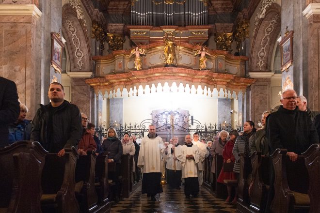 Wejście procesji do kościoła. Wierni stoją w ławkach, niektórzy ze złożonymi dłońmi. Nad wejściem widoczny chór z organami i złota figura świętego trzymającego laskę pasterską