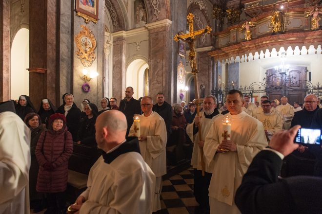 Procesja duchownych i zakonników idących przez nawę kościoła. Jeden z nich niesie wysoki krzyż, inni zapalone świece. W tle siostry zakonne i wierni w skupieniu obserwują wydarzenie