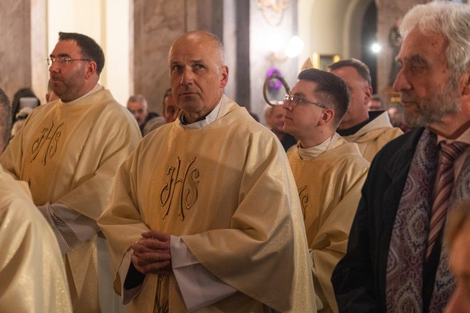 Zbliżenie na uczestników procesji. Starszy kapłan o poważnym wyrazie twarzy idzie wśród innych duchownych ubranych w kremowe ornaty. W tle tłum wiernych