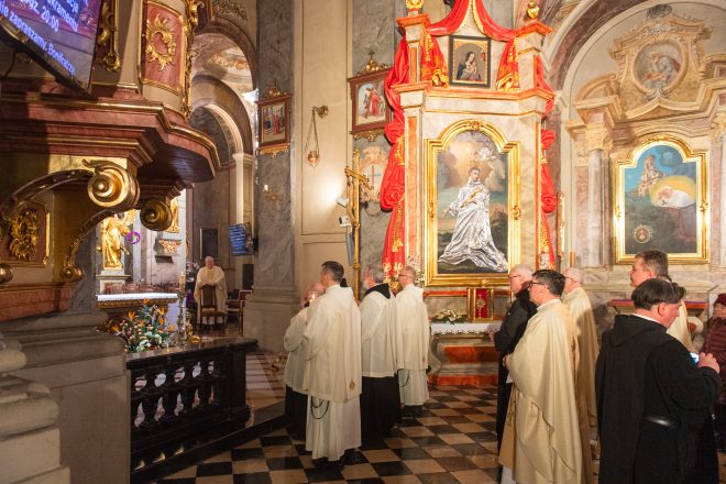 Kapłani i zakonnicy zgromadzeni przed bocznym ołtarzem w kościele. Centralnie wyeksponowany obraz świętego w złotej ramie, podświetlony ciepłym światłem