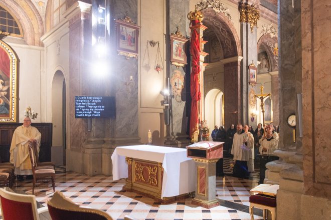 Widok na boczny ołtarz z białym obrusem. W tle tekst pieśni liturgicznej wyświetlany na ekranie. Wierni i duchowni wchodzą do głównej części świątyni, niosąc krzyż procesyjny