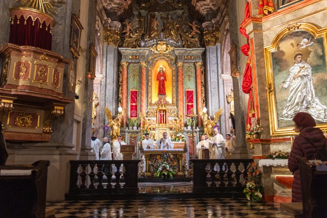 Główna część ceremonii liturgicznej. Kapłani odprawiają mszę przy bogato zdobionym ołtarzu, otoczeni złoconymi figurami i barokowymi dekoracjami. Wierni w skupieniu uczestniczą w modlitwie