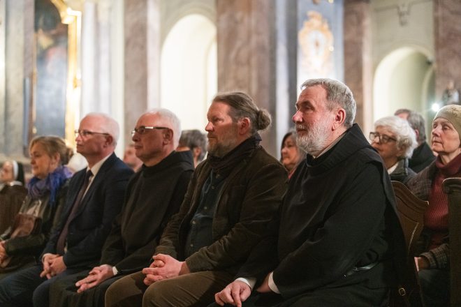 Grupa osób siedzących w ławkach zabytkowego kościoła. W pierwszym rzędzie widoczni są duchowni i świeccy uczestnicy ceremonii. W tle marmurowe kolumny i złocone zdobienia świątyni