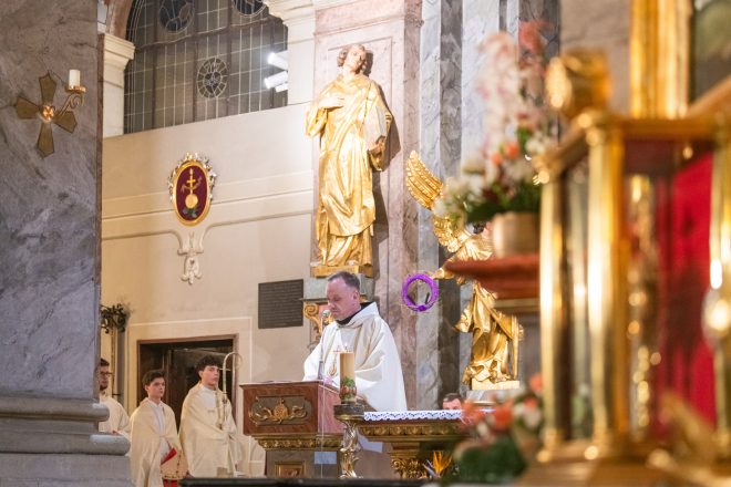 Duchowny w białym habicie wygłasza kazanie, stojąc przy ambonie. W tle złocona figura świętego oraz młodzi ministranci ubrani w białe alby