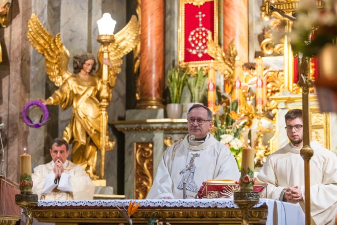 Kapłan ubrany w białą szatę liturgiczną stoi przy ołtarzu, wygłaszając modlitwę. Towarzyszą mu inni duchowni, a w tle widoczny jest bogato zdobiony ołtarz z kwiatami i złoconymi detalami