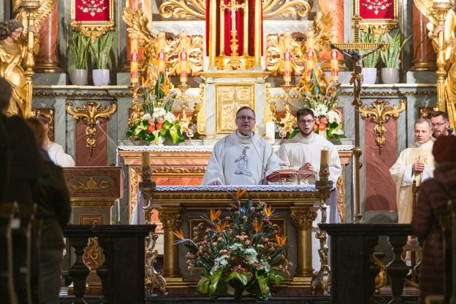 Celebrans w białej szacie liturgicznej stoi za ołtarzem, przemawiając do wiernych. Po bokach asystują duchowni, a w tle widoczna bogata dekoracja świątyni