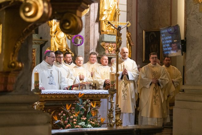 Kapłani ubrani w złote ornaty zgromadzeni wokół ołtarza podczas uroczystej mszy świętej. Jeden z duchownych trzyma duży procesyjny krzyż. W tle ekran z wyświetlonym tekstem modlitwy