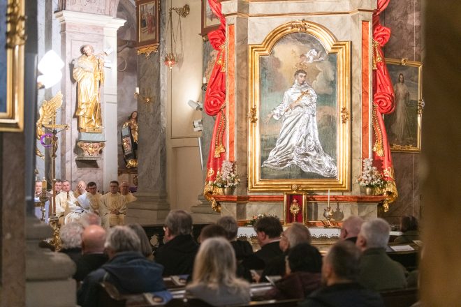 Fragment wnętrza kościoła z wiernymi siedzącymi w ławkach. Na pierwszym planie duży obraz świętego w złotej ramie, ozdobiony czerwoną tkaniną